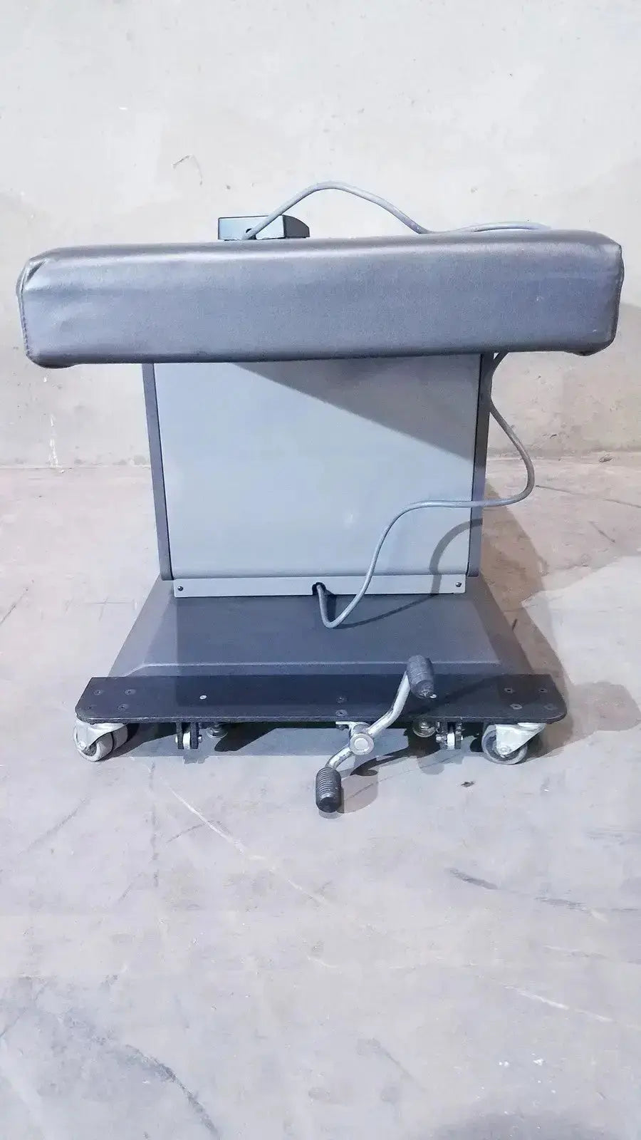 Gray padded headrest on metal frame with caster wheels and adjustable foot paddle of ritter 106 exam table hospital surplus