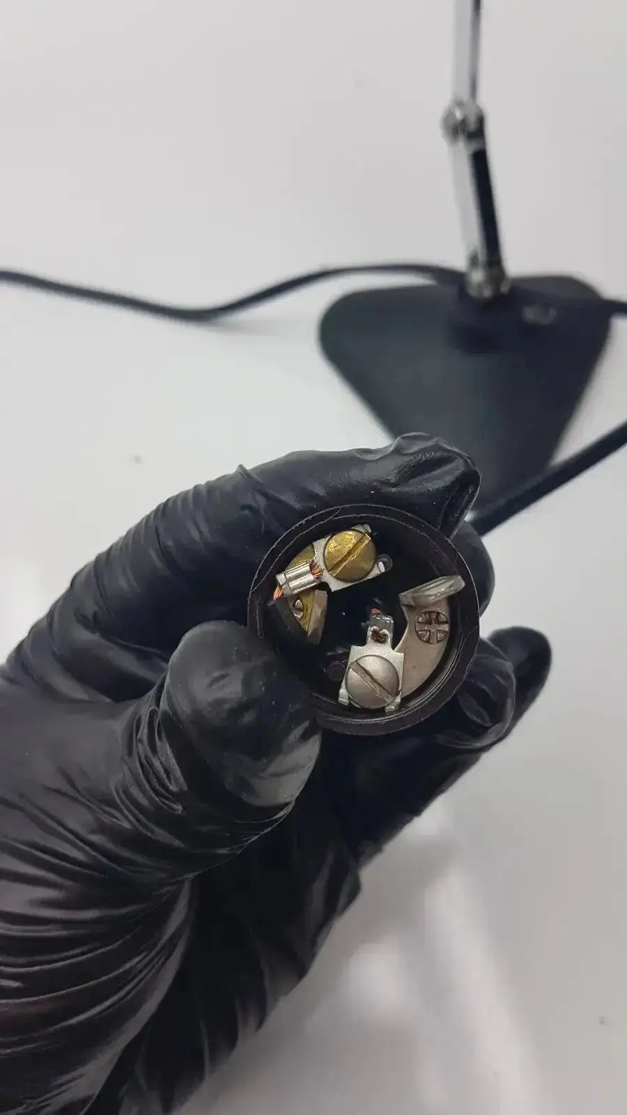 Black-gloved hand holding a circular metallic adjustable microscope illuminator light lamp with gold and silver components