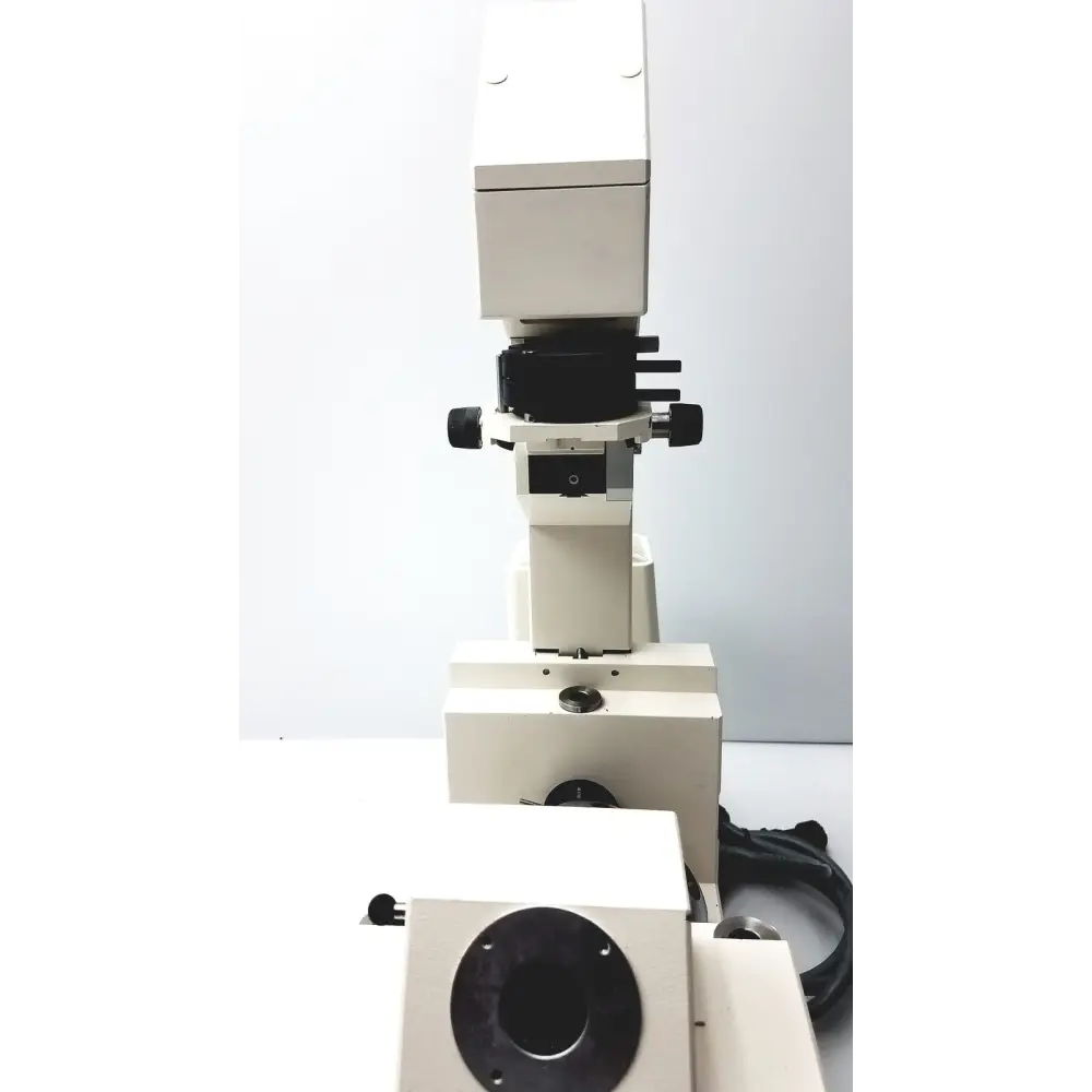 White and black compound microscope with a sturdy metallic base, featuring adjustable focus knobs and an objective lens turret.