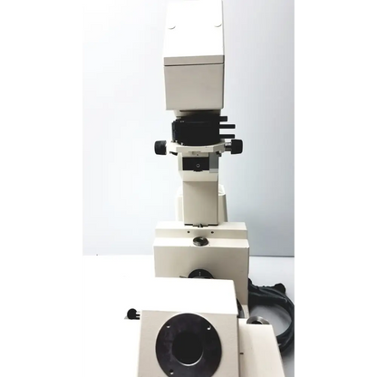 White and black compound microscope with a sturdy metallic base, featuring adjustable focus knobs and an objective lens turret.
