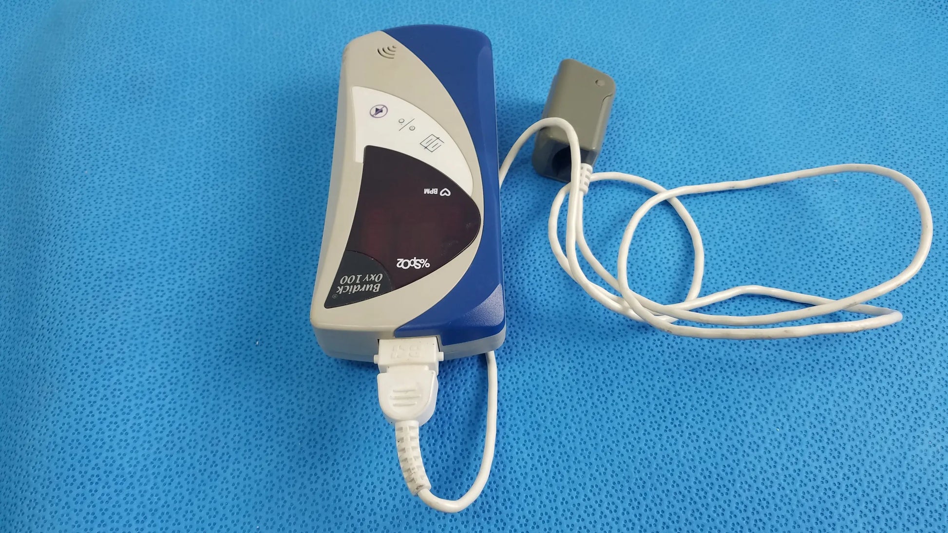 Blue and gray bionik ort-100 medical equipment with usb cable connected showing back side of burdick oxy 100 pulse oximeter ref 92521