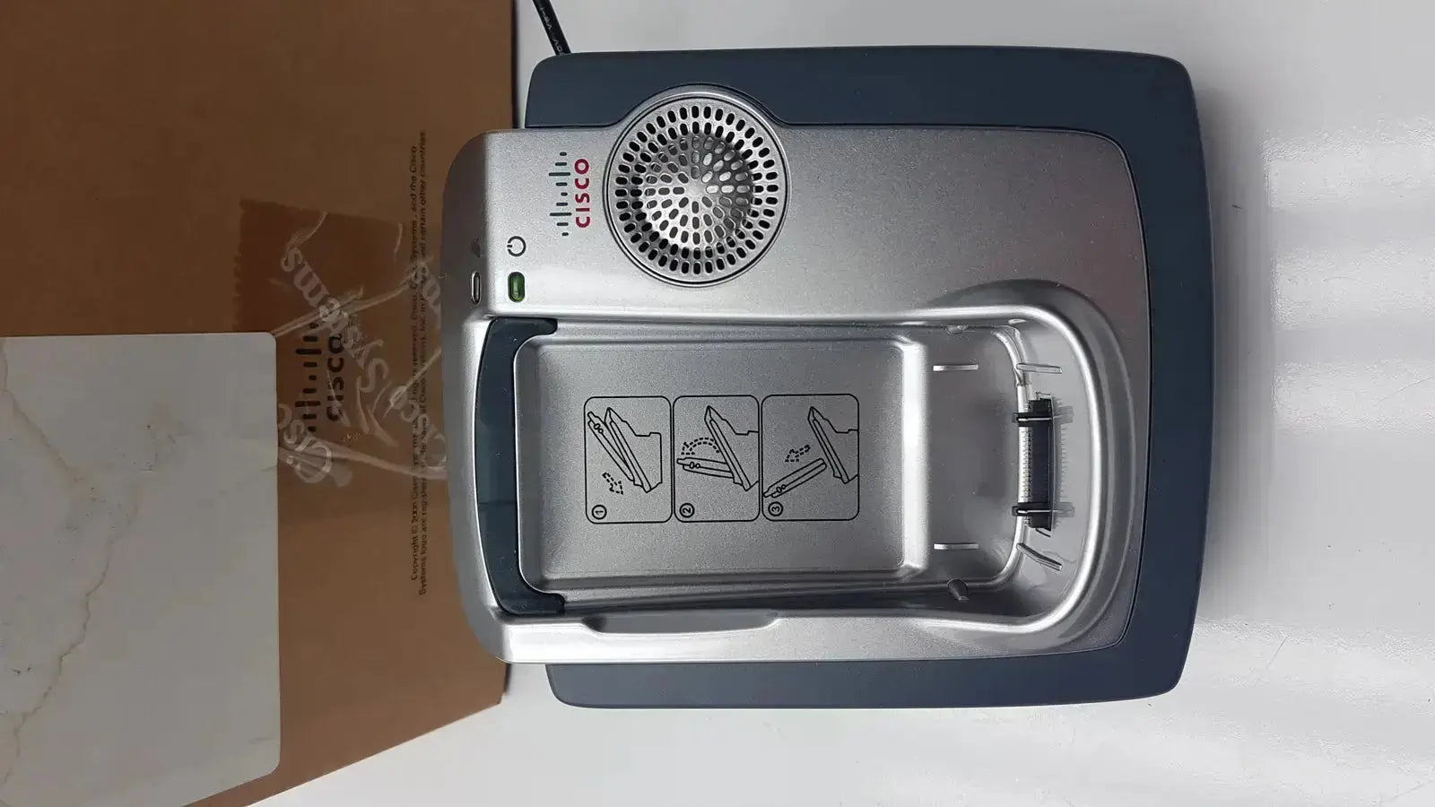 Silver cisco cp-7921g-a-k9 wireless phone with base featuring a circular speaker grille and instructional diagrams on front panel
