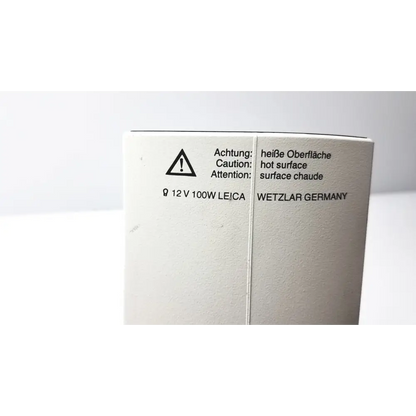 White cardboard box with black printed text and symbols, including a warning triangle for hot surface, voltage specification (12 v 100w), brand name ’leica,’ and location ’wetzlar germany.’