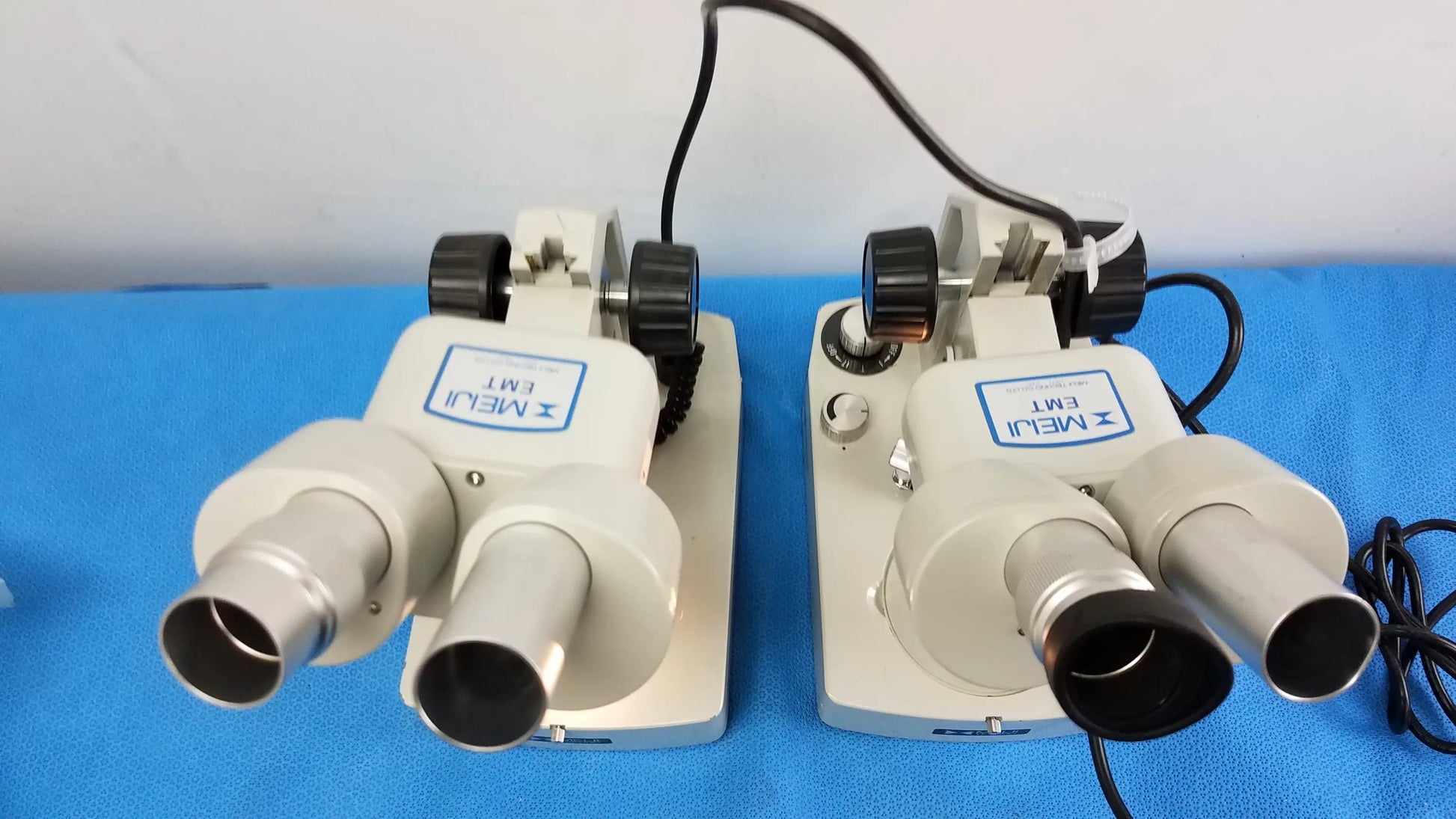 Two beige and silver meiji techno emt-1 stereo microscopes with black knobs on blue surface medical equipment