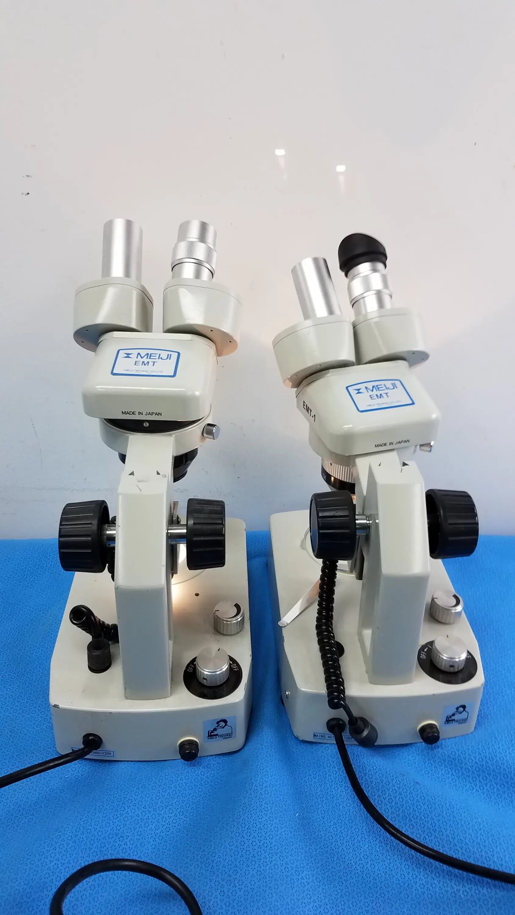 Two beige meiji techno emt-1 stereo microscopes with black knobs and silver eyepieces on blue surface medical equipment