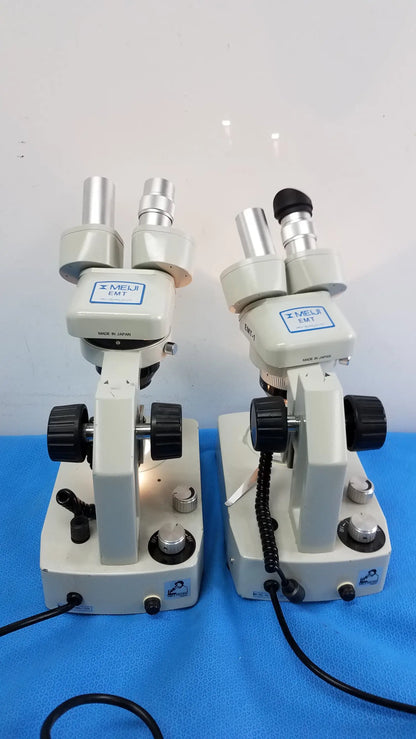 Two beige meiji techno emt-1 stereo microscopes with black knobs and silver eyepieces on blue surface medical equipment