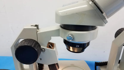 A white meiji techno stereo microscope with a black 2x objective lens and blue adjustment knob for medical equipment use