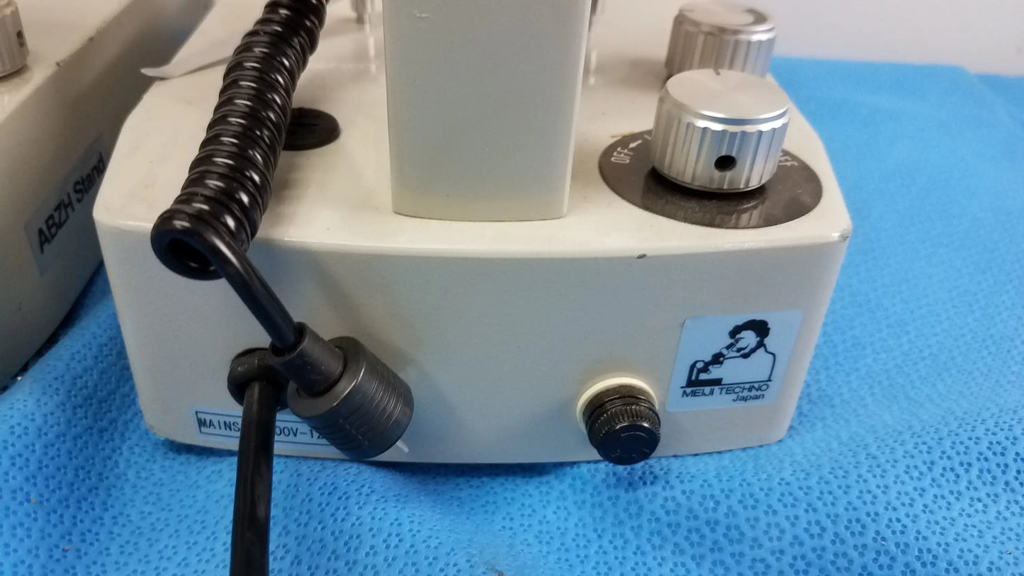 White rectangular Meiji Techno stereo microscope base with silver knobs and black coiled cable for medical equipment use