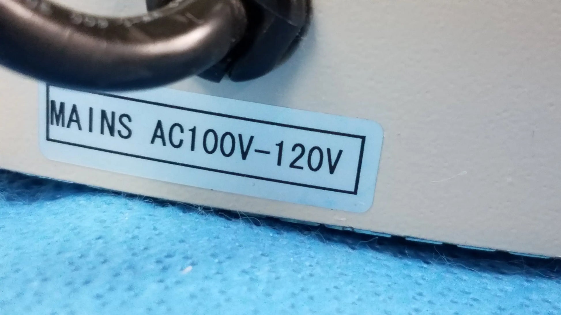 White rectangular label with black text mains ac100v-120v on the back of a beige inverted binocular stereo microscope