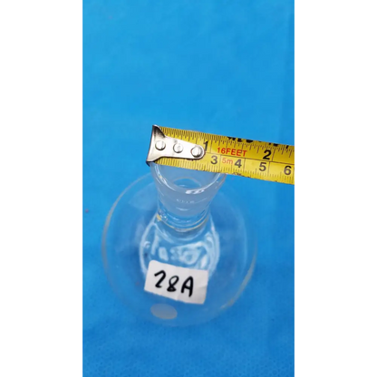 Clear glass erlenmeyer flask with a white label marked ’28a’ and a yellow measuring tape resting across its neck.