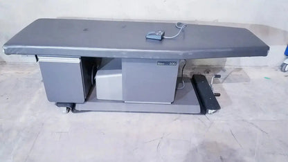 Gray metal ritter 106 exam table with padded surface and up and down foot paddle on wheeled hospital surplus base