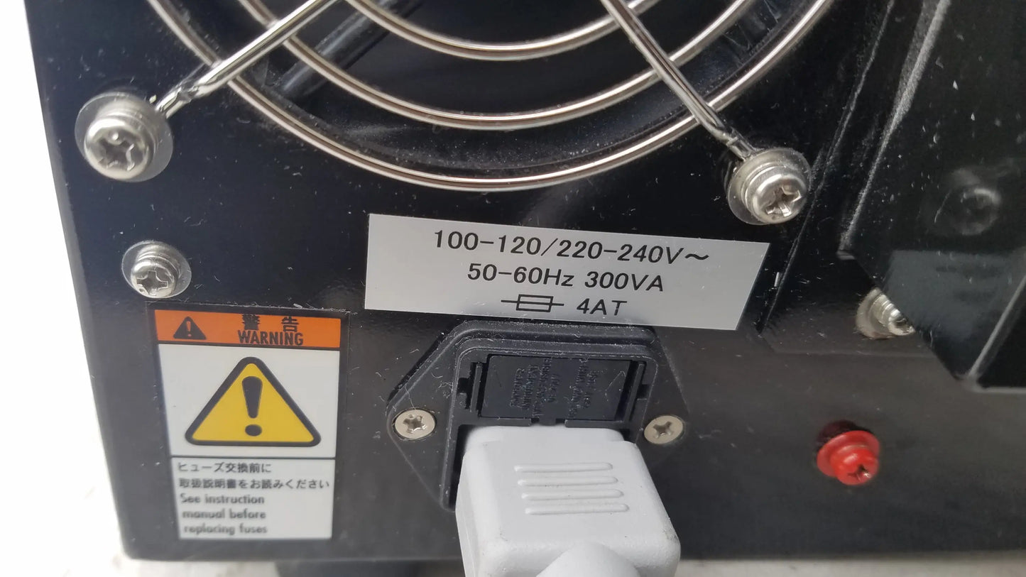 Black power supply unit with silver fan grille and white plug inserted showing voltage specs on Shimadzu MCE-202 medical equipment back side
