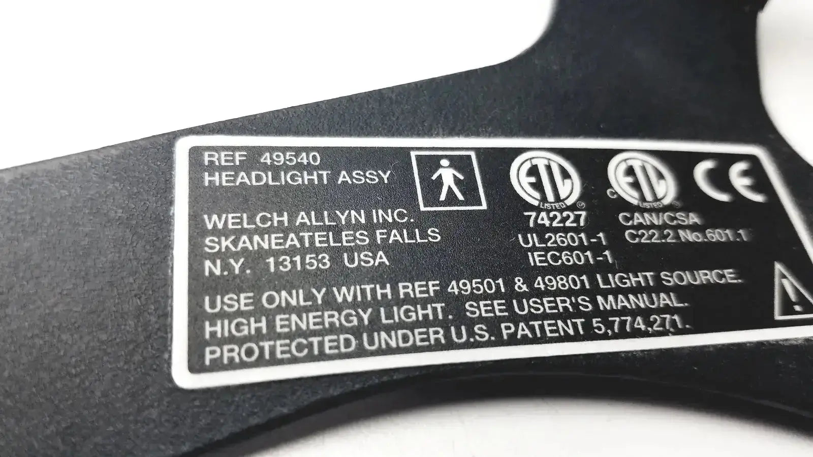Black plastic label with white text and certification marks on the back of welch allyn 49540 surgical headlight assy